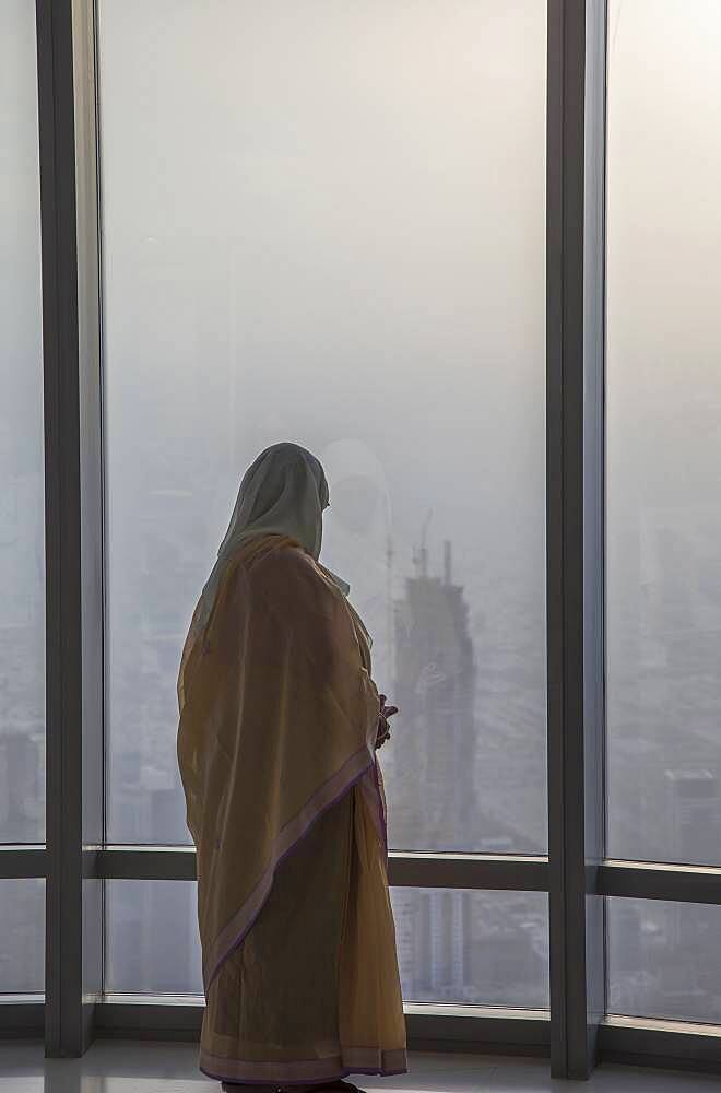 Asian woman at window admiring Dubai cityscape, Dubai Emirate, United Arab Emirates