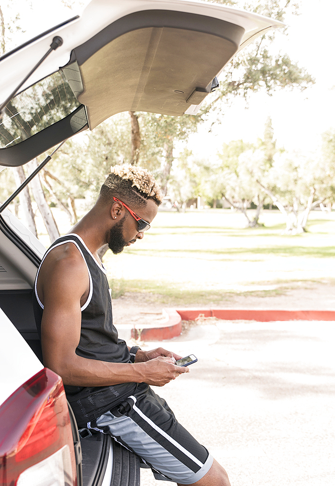 Black man leaning on car hatchback texting on cell phone