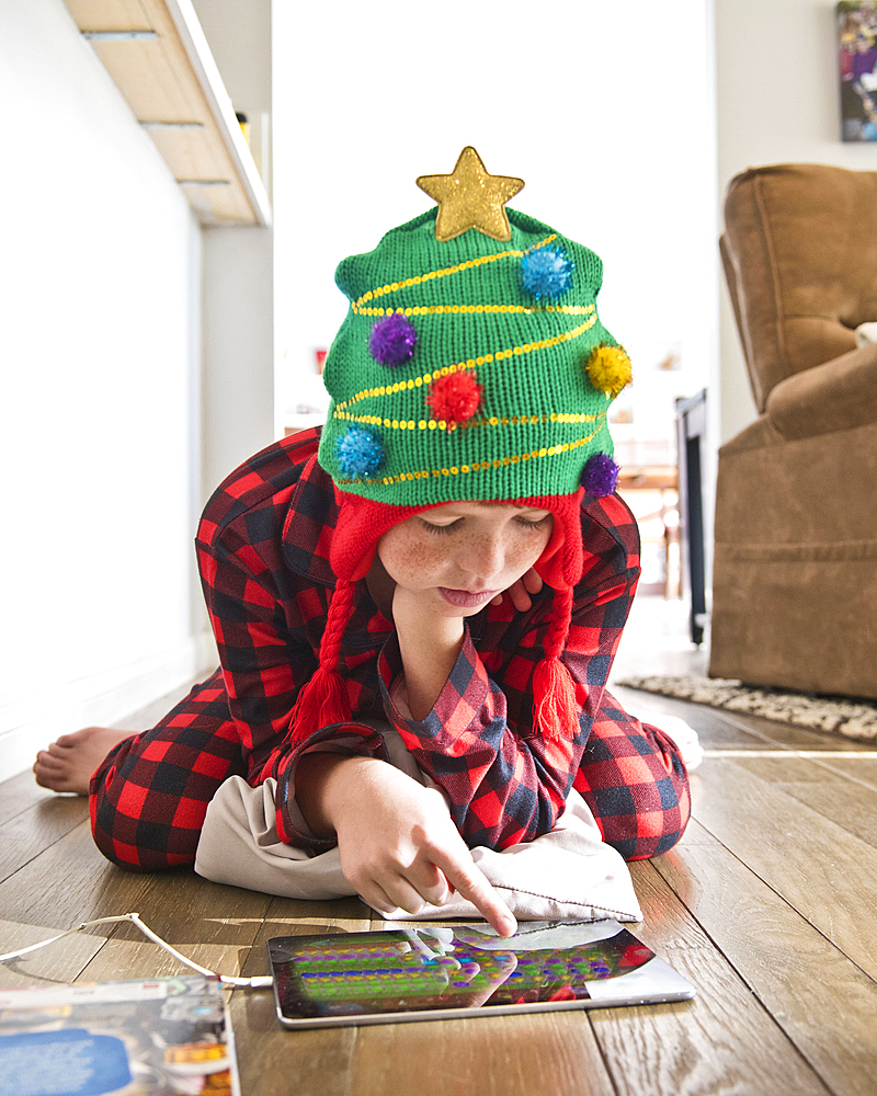 Caucasian boy wearing Christmas tree stocking-cap using digital tablet on floor