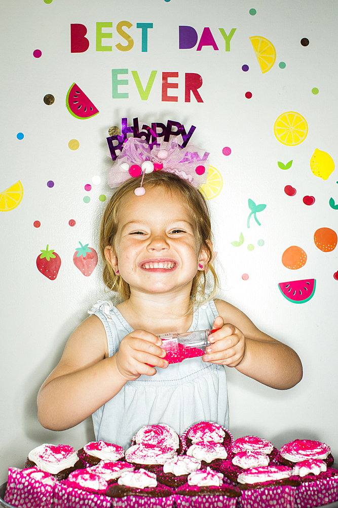 Caucasian girl holding sprinkles over tray of cupcakes for birthday