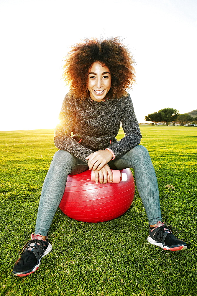 Smiling Hispanic woman resting on fitness ball in sunny field