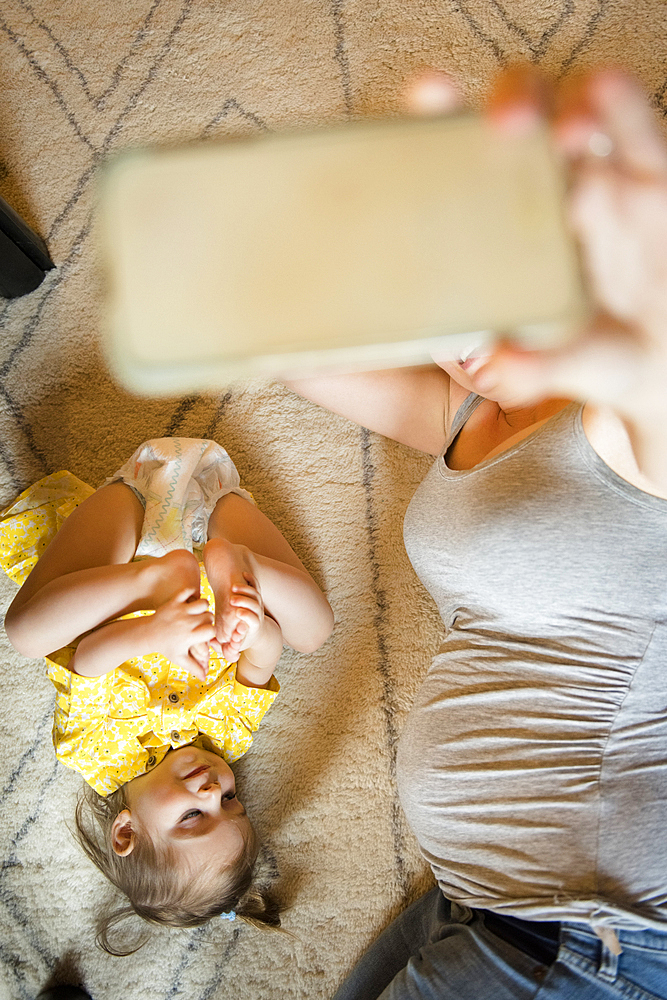 Expectant mother taking cell phone selfie on floor with Caucasian daughter