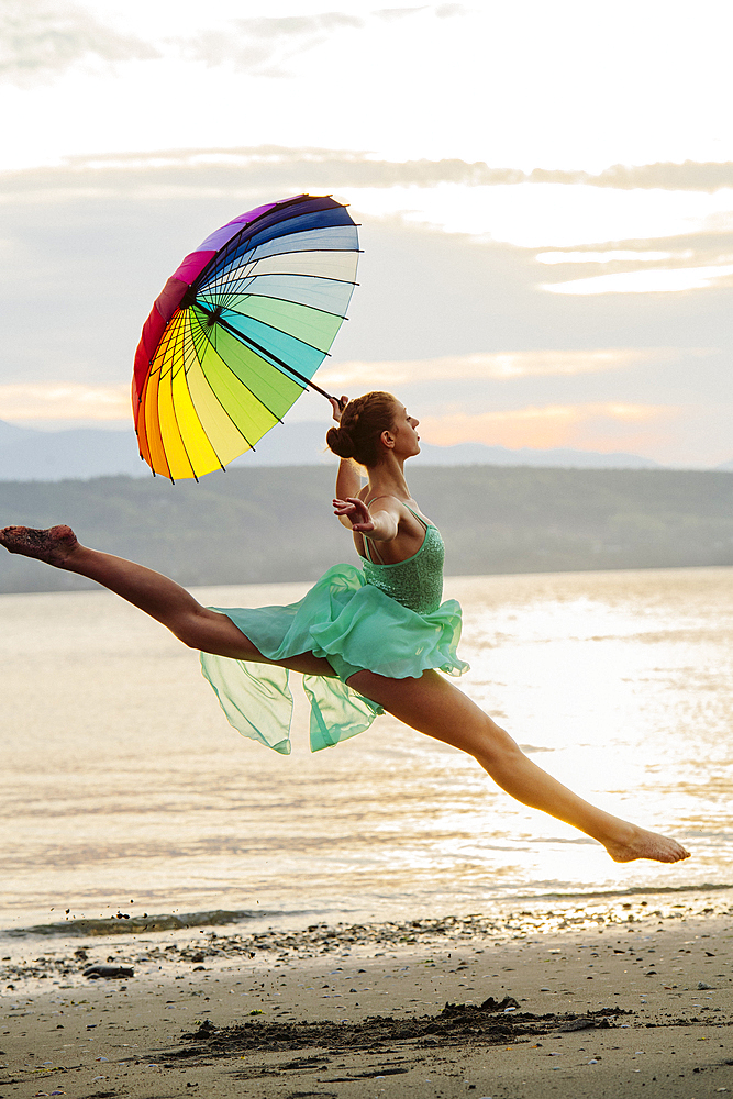 Caucasian ballerina jumping with multicolor umbrella on beach