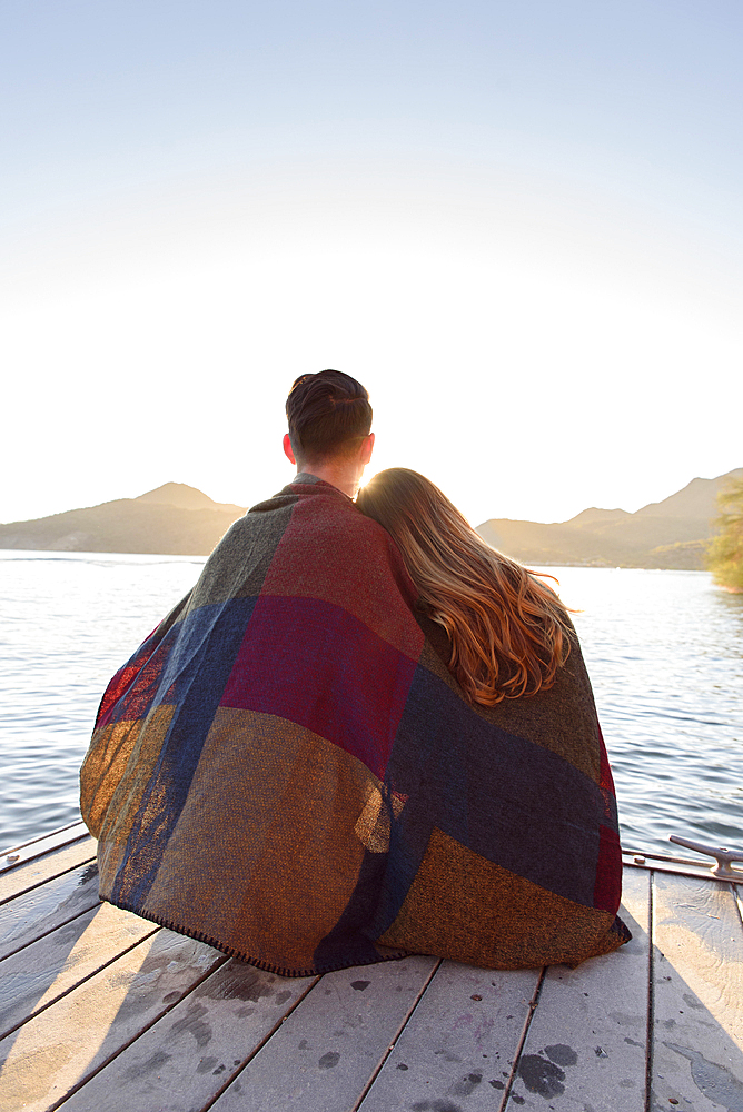 Couple sitting on dock at river wrapped in blanket