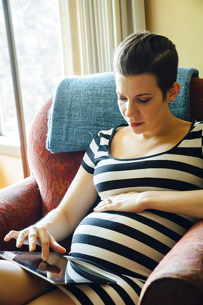 Pregnant Caucasian woman sitting in armchair using digital tablet