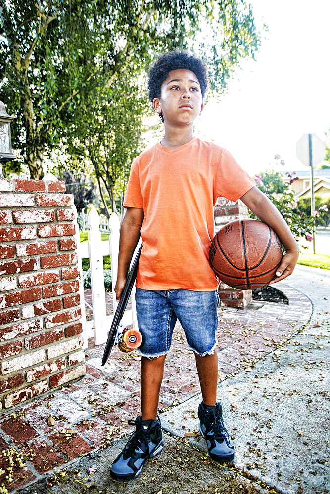 Mixed Race boy standing on sidewalk holding skateboard and basketball