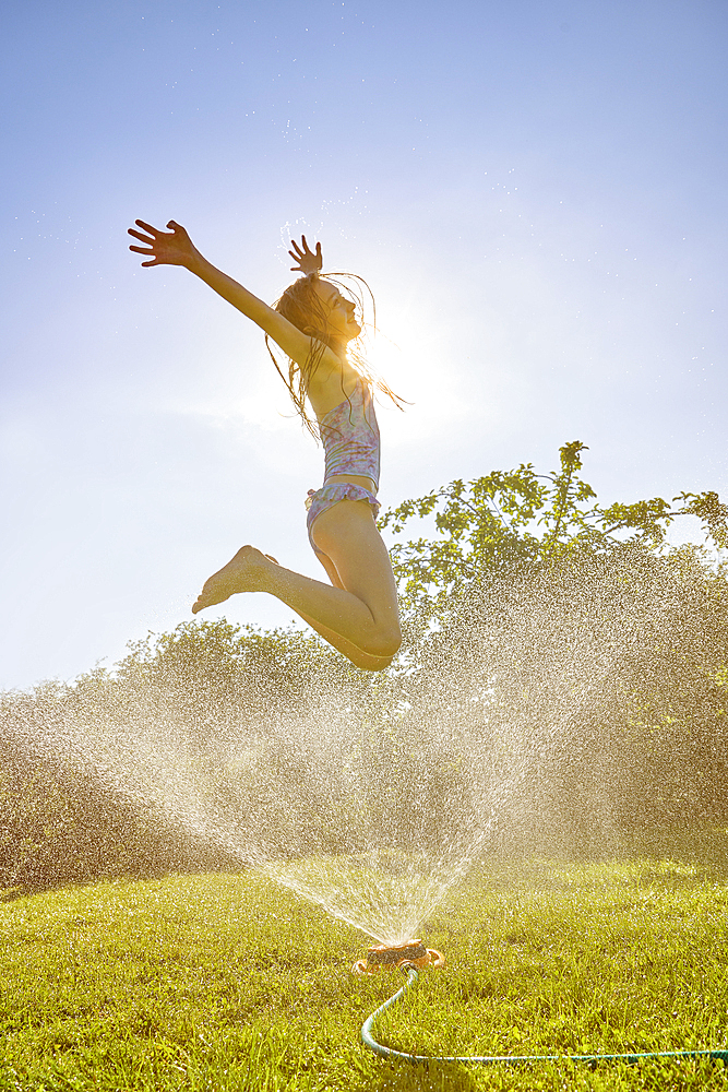 Caucasian girl jumping over backyard sprinkler