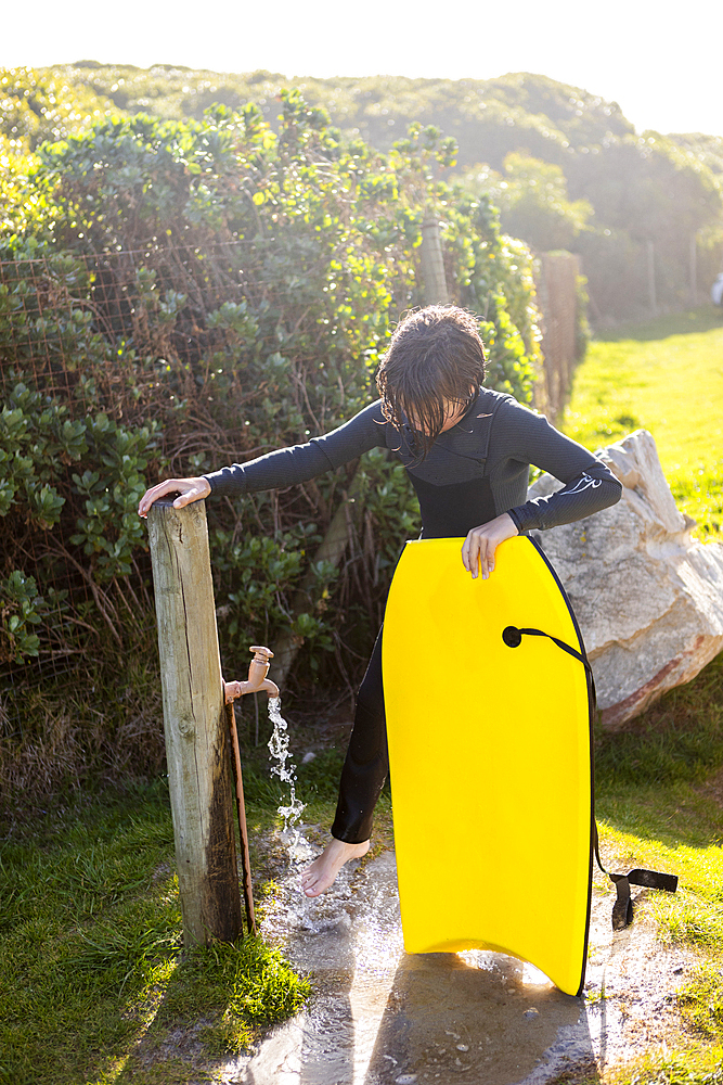 Boy (10-11) with surfboard using outdoor faucet on Kammabaai Beach