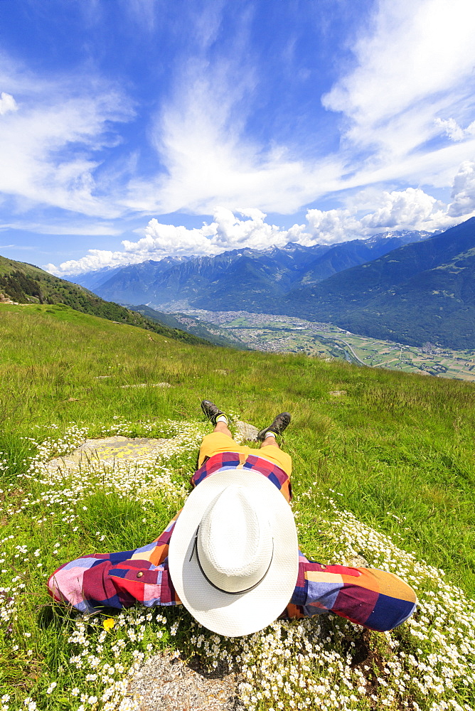 Rear view of man with hat lying on grass, Alpe Bassetta, Valtellina, Sondrio, Lombardy, Italy, Europe