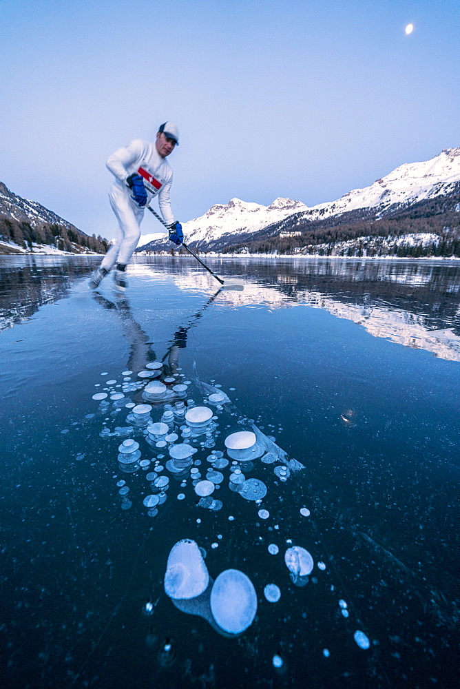 Ice hockey player man skating on Lake Sils covered in  ice bubbles at dusk, Engadine, Graubunden canton, Switzerland, Europe