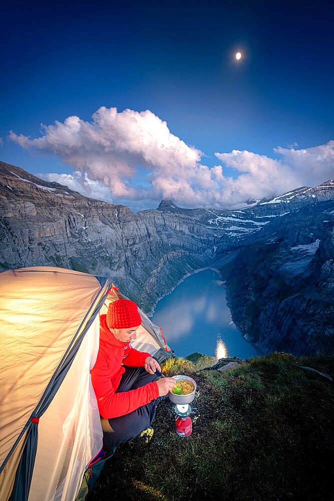 Hiker man cooking while camping in a tent above lake Limmernsee, Canton of Glarus, Switzerland, Europe