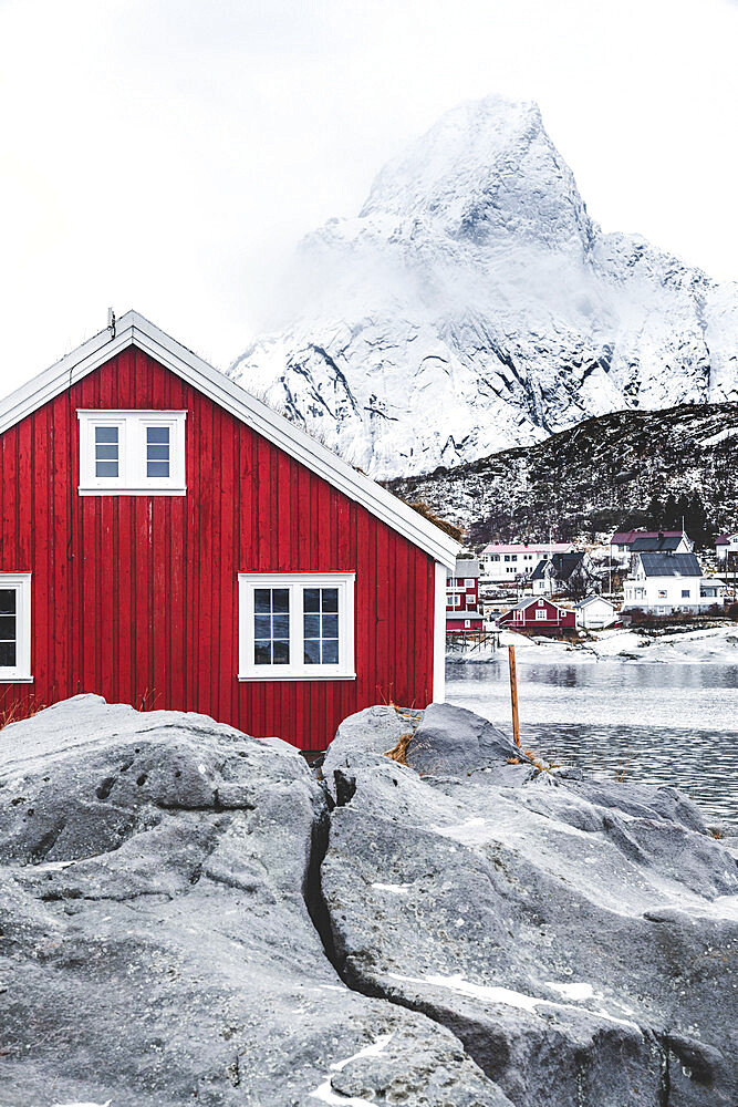 Red Rorbu in the frozen landscape with snowcapped Olstind mountain in the background, Reine, Nordland, Lofoten Islands, Norway, Scandinavia, Europe