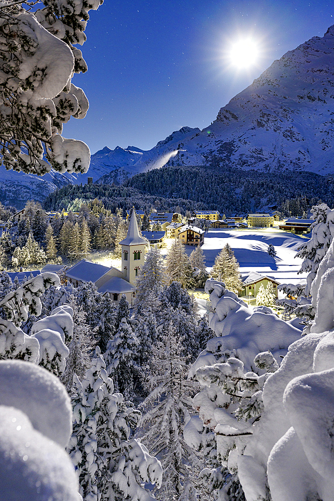 Full moon over Chiesa Bianca covered with snow surrounded by woods, Maloja, Bregaglia Valley, Graubunden, Engadin, Switzerland