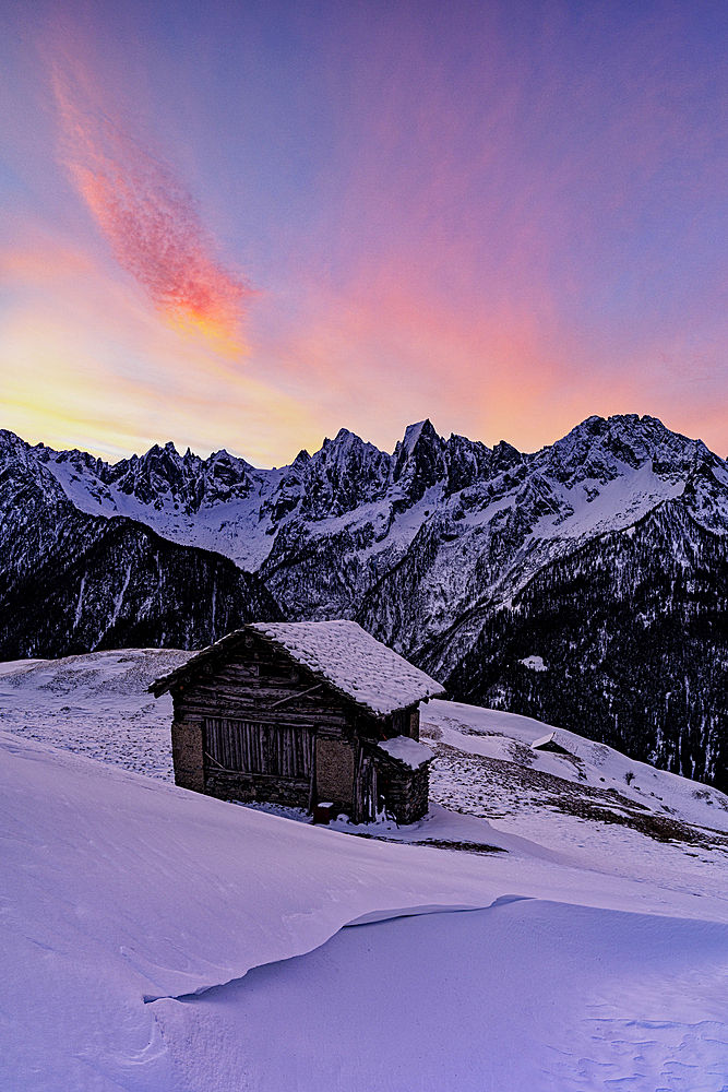 Lone mountain hut in deep snow with majestic peaks in the background at dawn, Tombal, Val Bregaglia, Graubunden, Switzerland, Europe