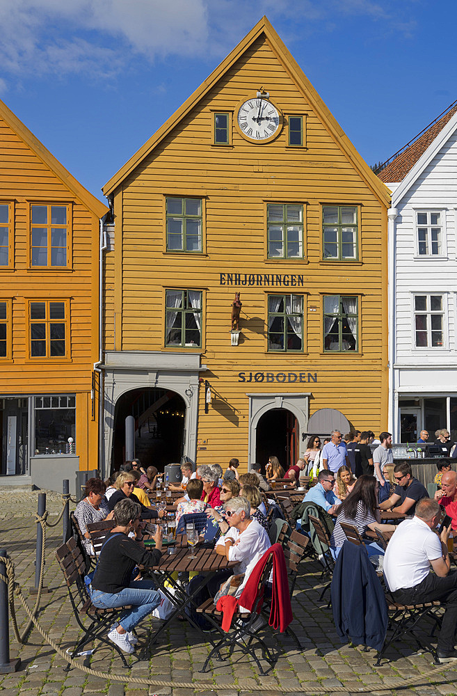 Tourists sit in the sun in outdoor bars and cafes in the old wharf and traditional wooden buildings in the Bryggen quarter of Bergen, Norway, Scandinavia, Europe