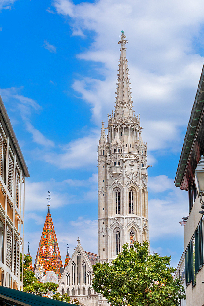 Street-level view of the Gothic Matthias Church tower and spire framed by nearby buildings in the Castle District. Vertical portrait orientation.