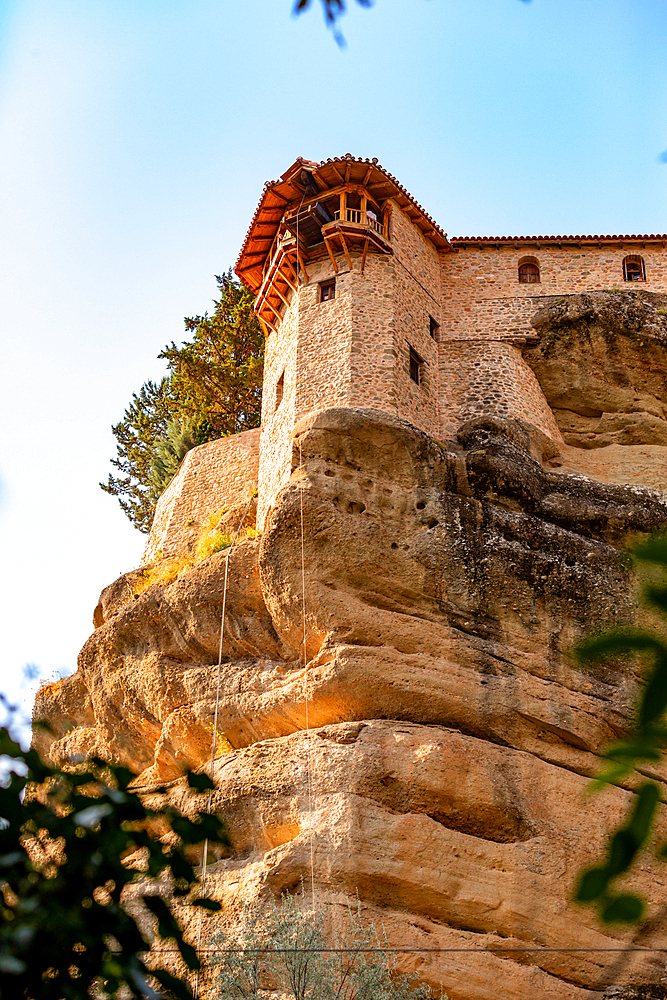 Vertical ground-level shot looking up at a stone monastery tower built on the edge of a steep rock cliff, with warm sunlight highlighting the textures and a soft blue sky above.
