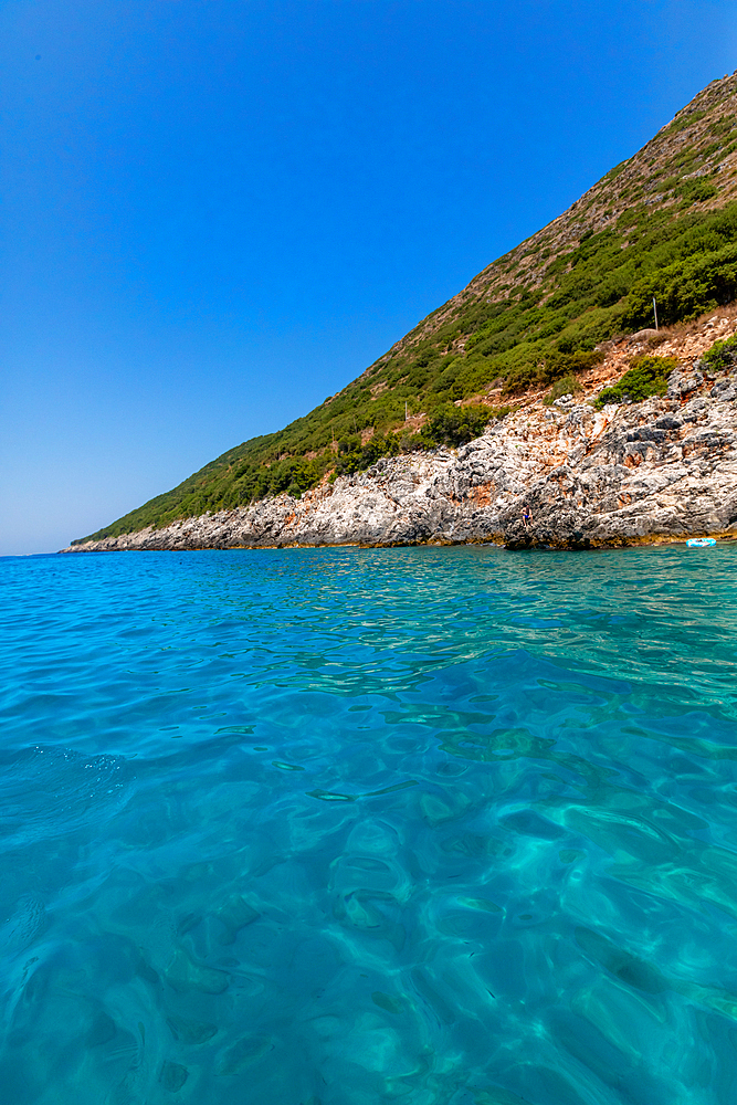 Clear turquoise waters of the Ionian Sea meeting rocky, vegetation-covered coastline near Dhermi, Vlorë County, Albania, on a bright summer day.
