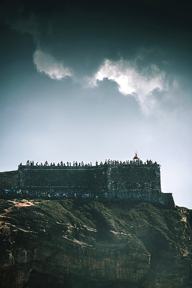 View of tourists at the iconic Nazare Lighthouse, Portugal on a partially cloudy day which overlooks the North Beach with some of the world's largest waves