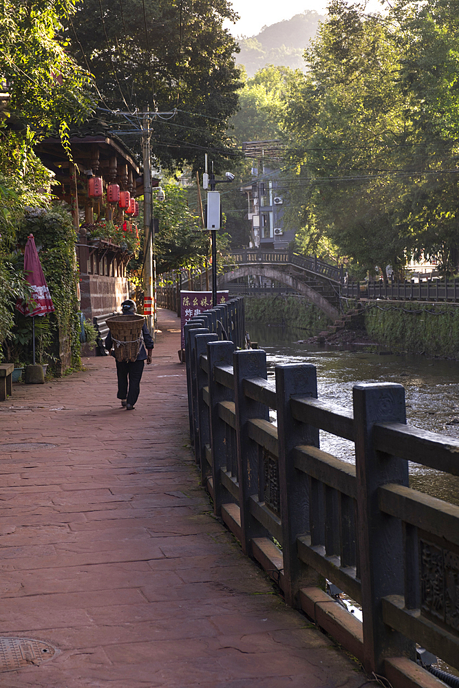 A woman with a wicker basket on his shoulders walking at dawn in a riverside path in Shagli town