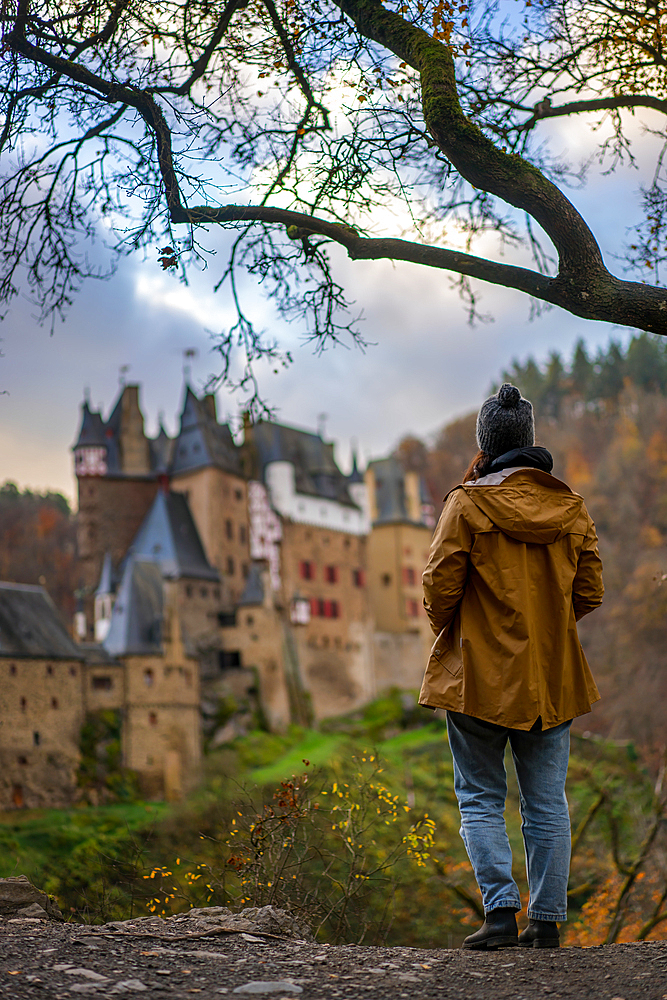 Back view of young woman with yellow jacket looking at Eltz medieval historic castle in an autumn landscape with trees at sunrise, Wierschem, Rhineland-Palatinate, Germany, Europe