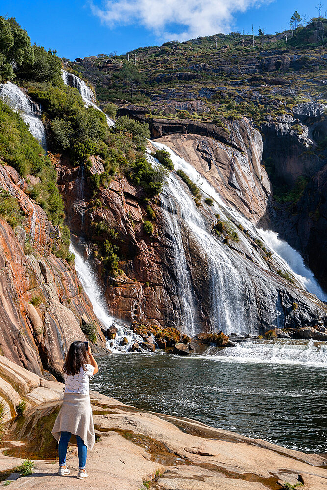 Caucasian woman photographing Ezaro waterfall with water crashing on lake between rocks in Galicia, Spain, Europe