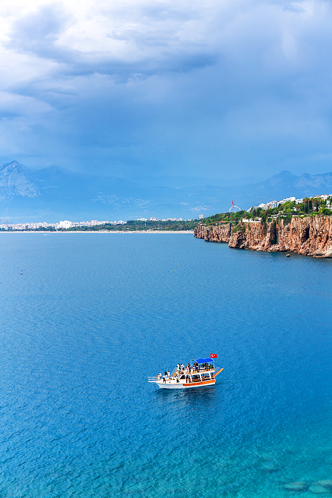 Antalya city view near the cliffs with a tourist boat on the sea with turquoise water and mountains in the background, in Turkey