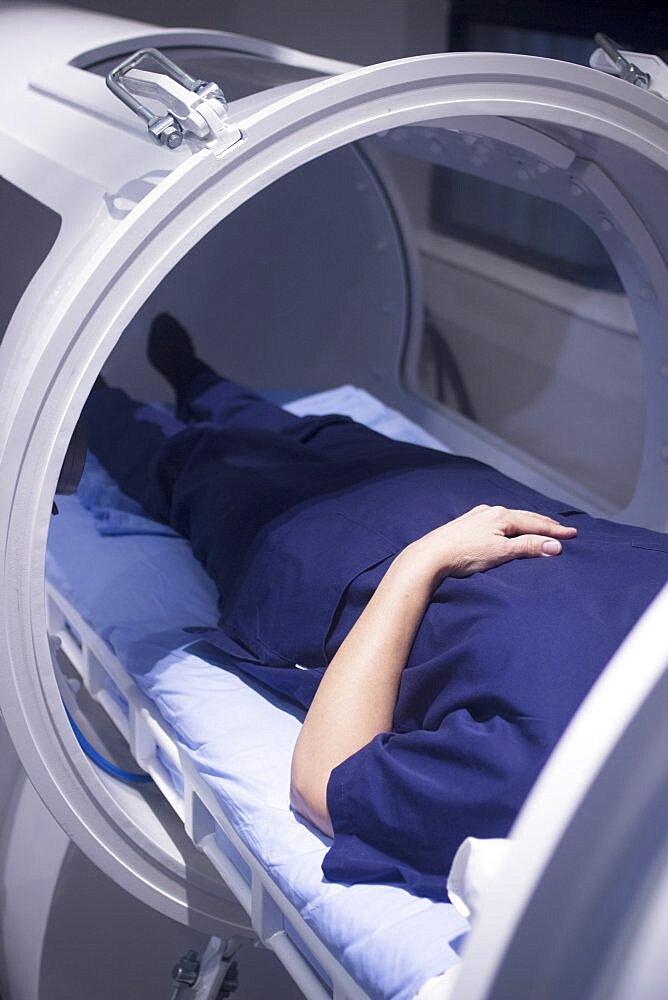 Female patient aged 45-55 wearing flower dress lying down in hyperbaric oxygen chamber receiving Hyperbaric Oxygen Therapy (HBOT) specialised medical treatment for injuries.