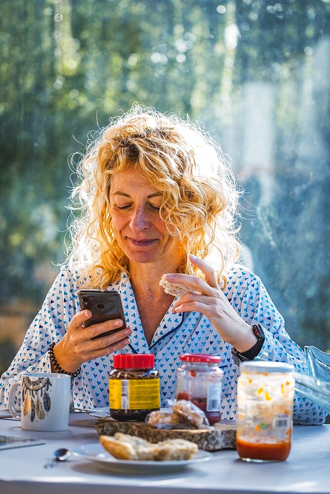 Blonde young mature woman in pyjamas at home in breakfast time looking his mobile and eating a toast.