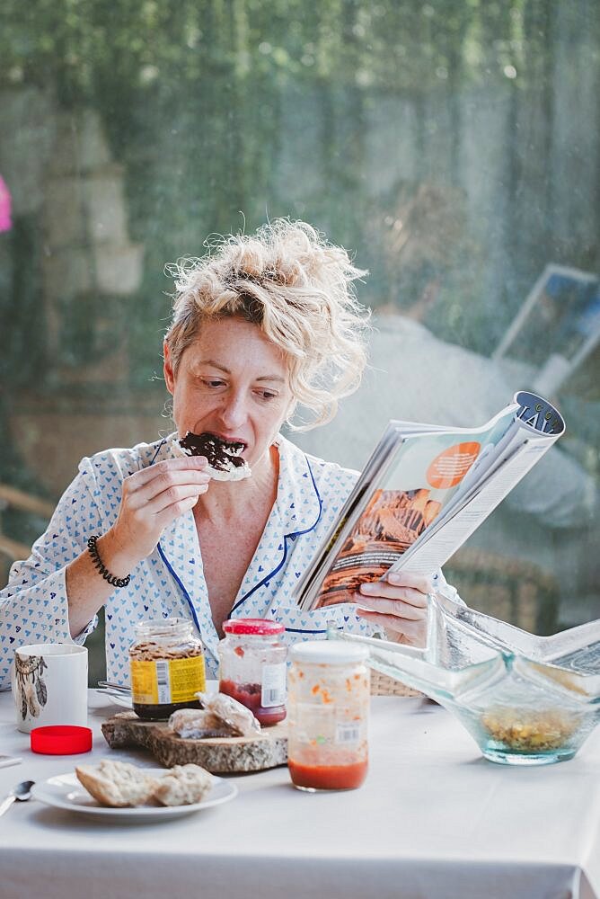 Blonde young mature woman in pyjamas at home in breakfast time, reading a magazine and eating a toast.