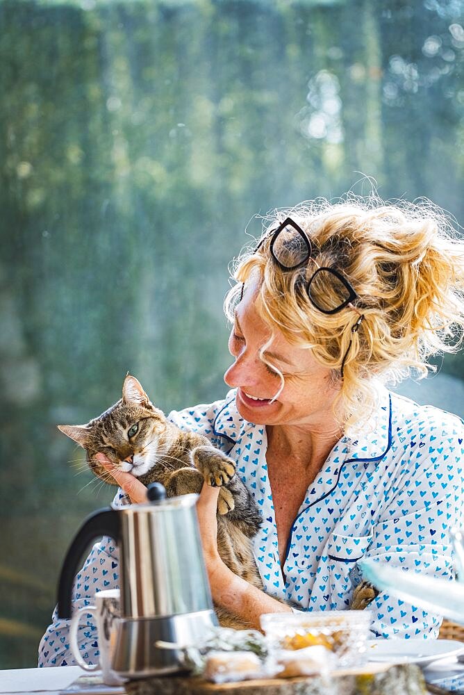 Blonde young mature woman with glasses in pijamas at home in breakfast time, reading a magazine and having a moment with her cat.