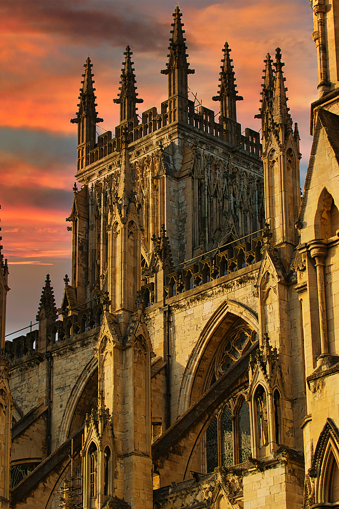 A close-up view of a stone cathedral against a pink and orange sunset sky. The image focuses on the intricately carved spires and arches, with visible stonework and details.