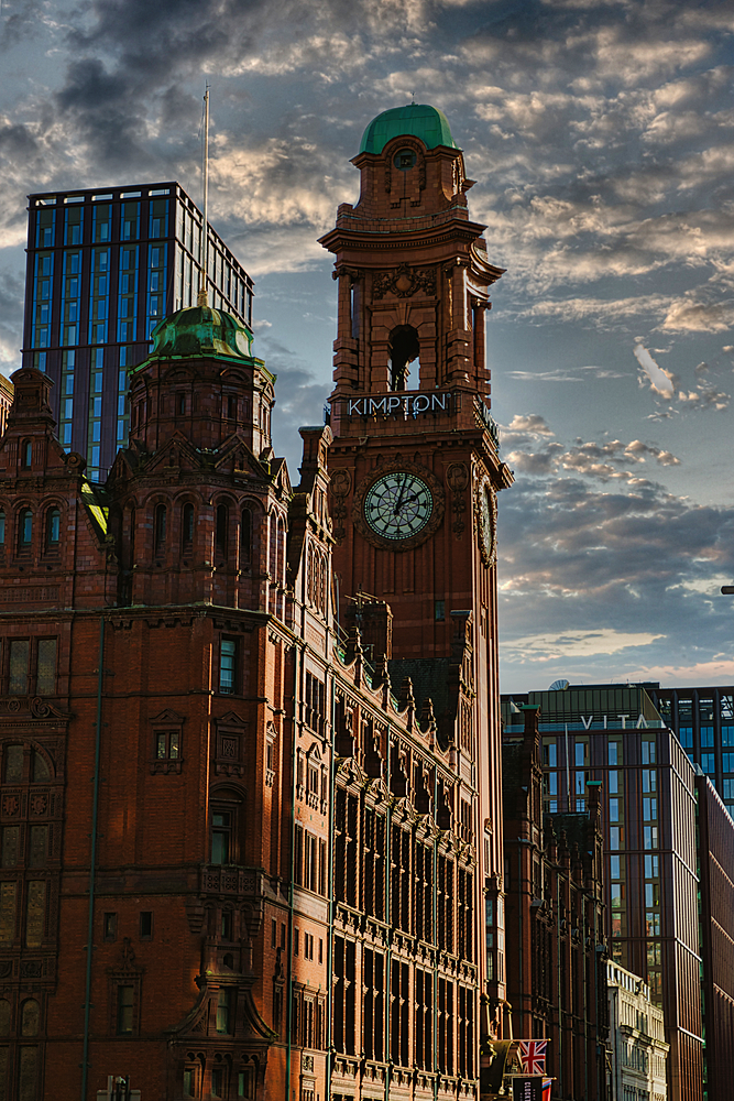 A tall, brick building with a clock tower and green roof, glass-fronted skyscraper under a cloudy sky in Manchester, UK.