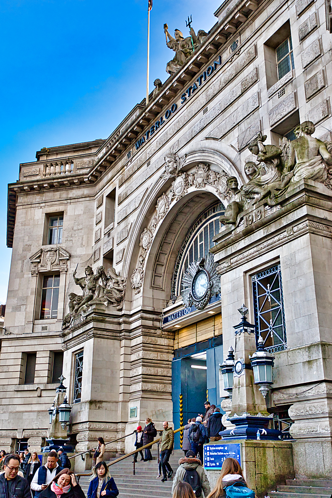 Exterior view of Waterloo Station's main entrance, showcasing its elaborate stone facade, statues, arched entryway, clock, and people ascending the steps. The building is predominantly light beige stone in London, UK.