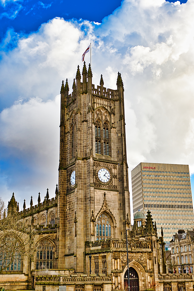 A tall, stone church with a prominent clock tower dominates the image, set against a partly cloudy sky. A modern office building is partially visible in the background in Manchester, UK.