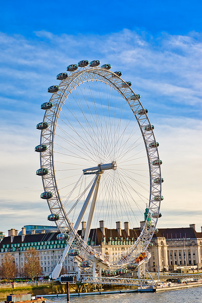 The London Eye Ferris wheel dominates the image, its capsules visible against a clear blue sky. Buildings line the riverbank in the background.