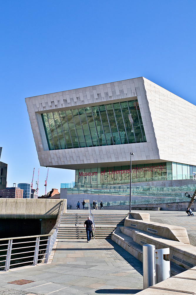 Museum of Liverpool with an angled facade, large glass windows, clear blue sky, and a man walking up the steps toward the entrance.
