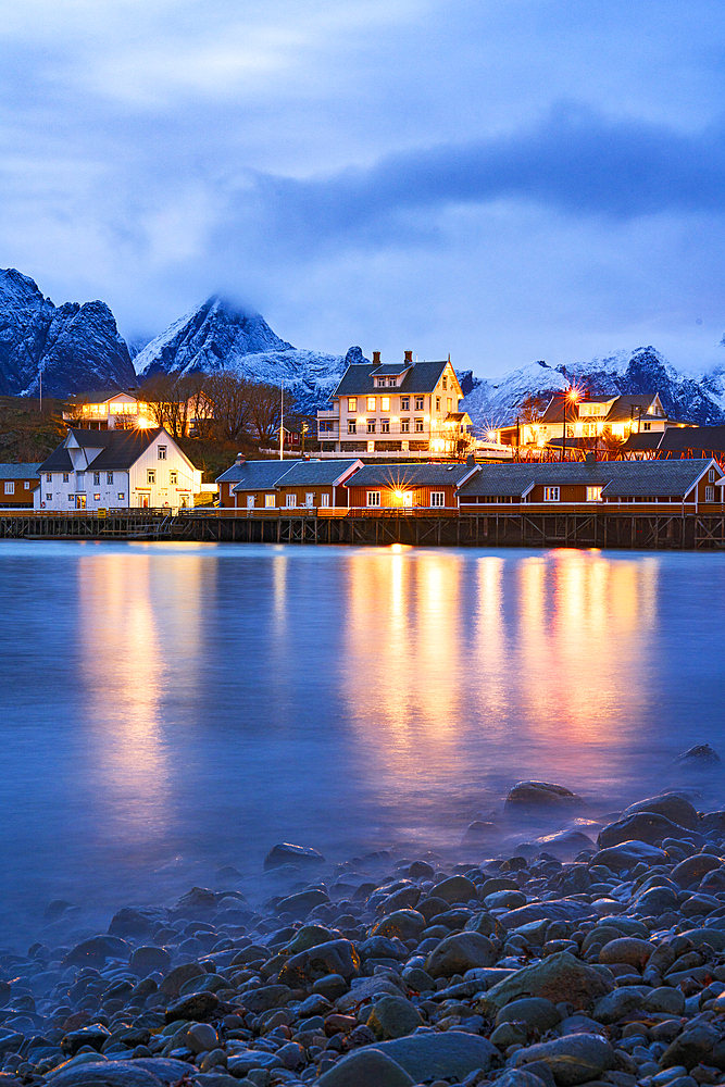 Illuminated rorbu reflected in the sea at dusk, Sakrisoy, Reine, Lofoten Islands, Norway