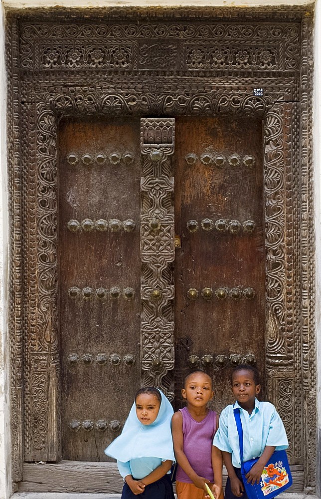 Three children sitting in front of a carved wooden Arab door in Stone Town, Zanzibar, Tanzania, East Africa