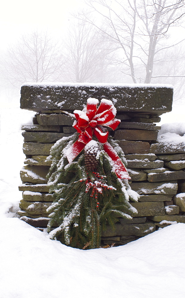 A snow covered Christmas wreath of pine branches, red berries and ribbon hanging on a stone wall, Rensselaervile, New York, United States of America, North America