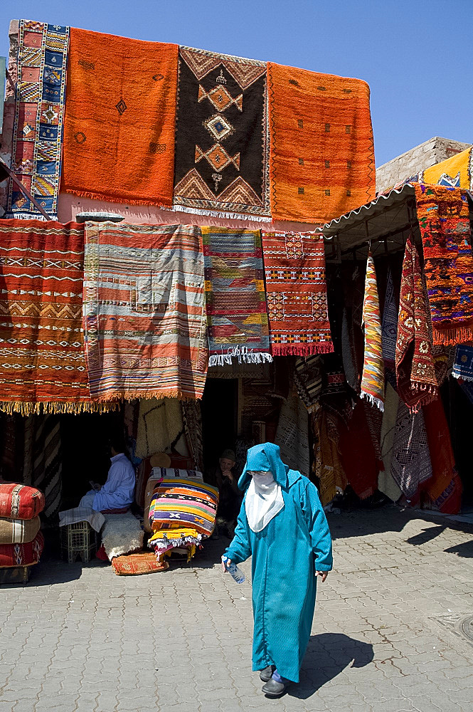 A woman in traditional Islamic dress walking past carpets hanging around an entrance to the souk in Marrakech, Morocco, North Africa, Africa