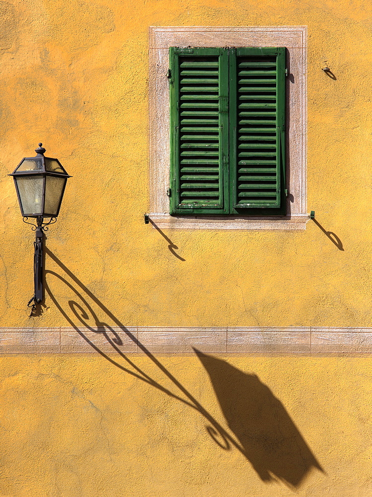 Architectural detail showing green wooden shutter on yellow wall with ornate wall lamp and shadow, San Quirico d'Orcia, Tuscany, Italy, Europe