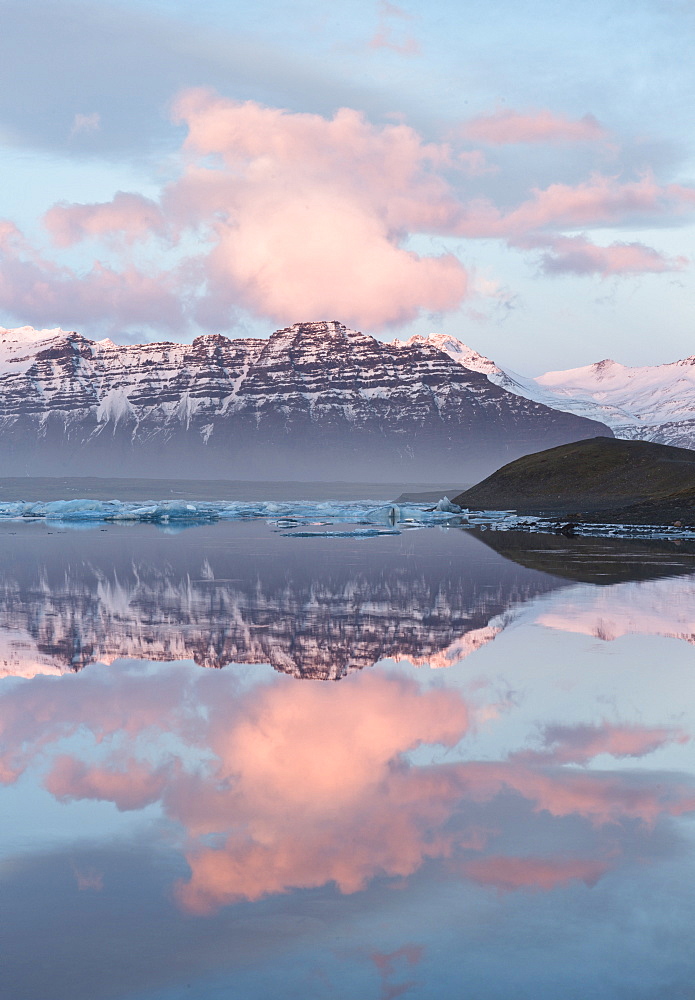 Panoramic view across the calm water of Jokulsarlon glacial lagoon towards snow-capped mountains and icebergs bathed in the last light of a winter's afternoon, at the head of the Breidamerkurjokull Glacier on the edge of the Vatnajokull National Park, South Iceland, Iceland, Polar Regions