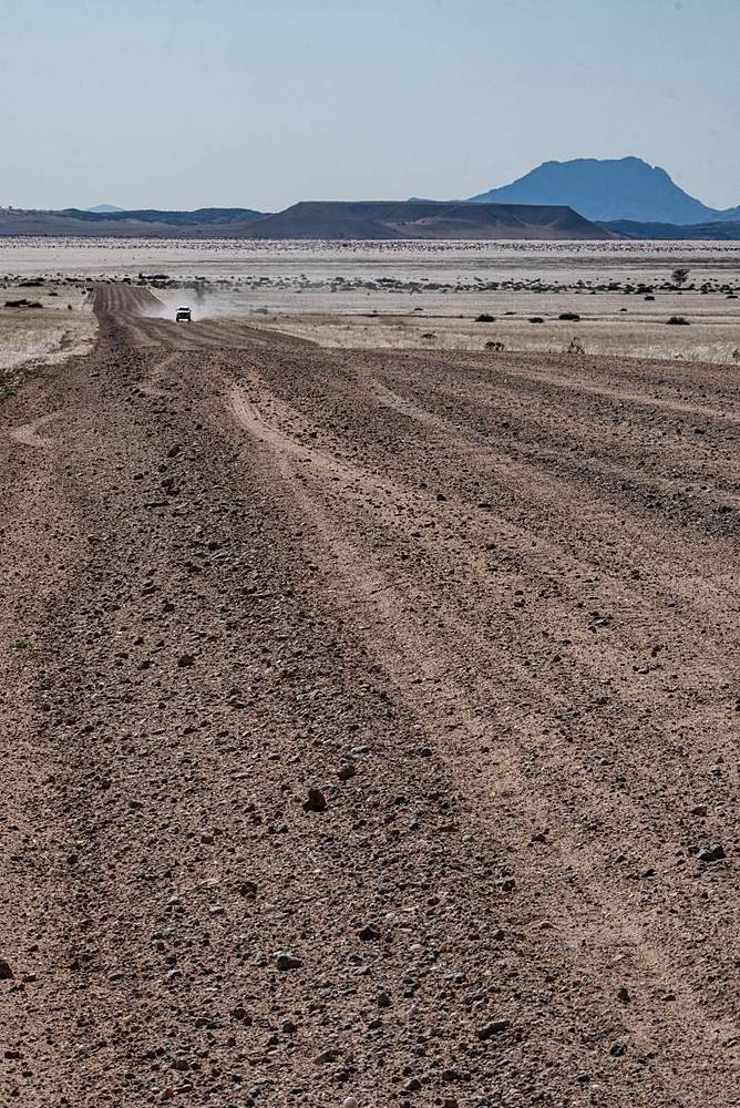 Four by four vehicle in the distance on long straight gravel road, blue mountains in the background, north of Solitaire, Namibia, Africa