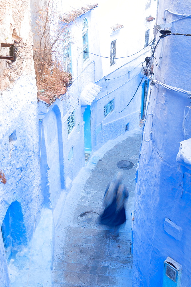 Woman in dark blue traditional clothing running down (blurred) a typical blue painted alleyway in Chefchaouen, Morocco, North Africa, Africa