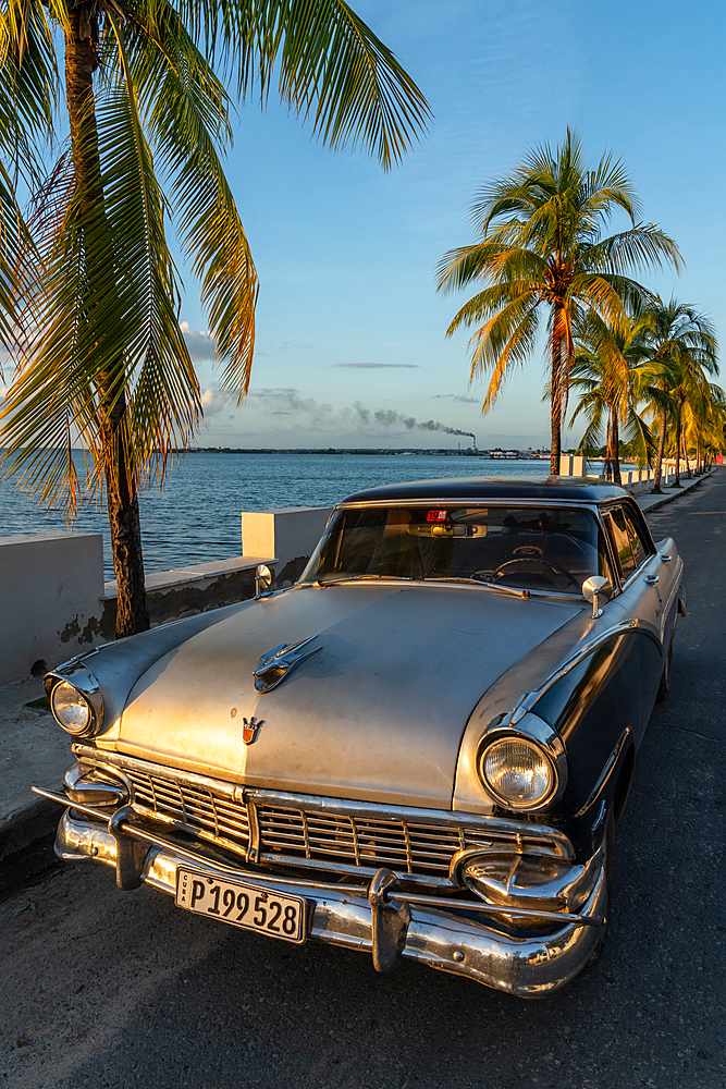 Classic silver Ford car parked on lonely coastal road, refinery in background, Cienfuegos, Cuba