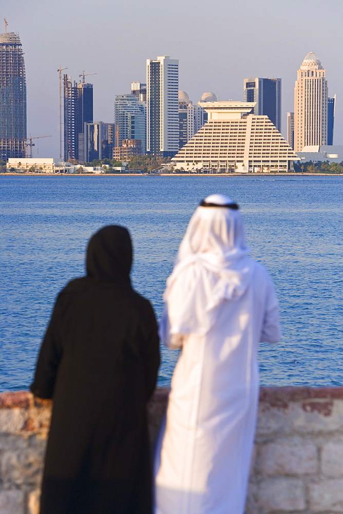 Man and woman in traditional dress looking across Doha Bay from the Corniche to the new city skyline and West Bay business and financial district, Doha, Qatar, Middle East