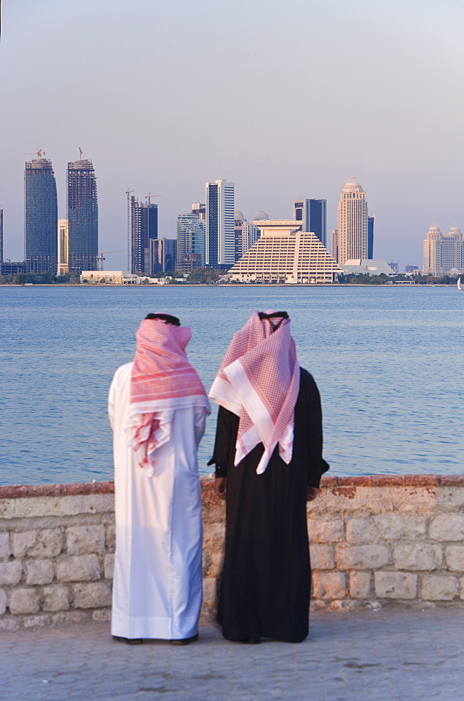 Two men wearing traditional dress of thobe and gutra (headdress) looking across Doha Bay from the Corniche to the new city skyline and West Bay business and financial district, Doha, Qatar, Middle East