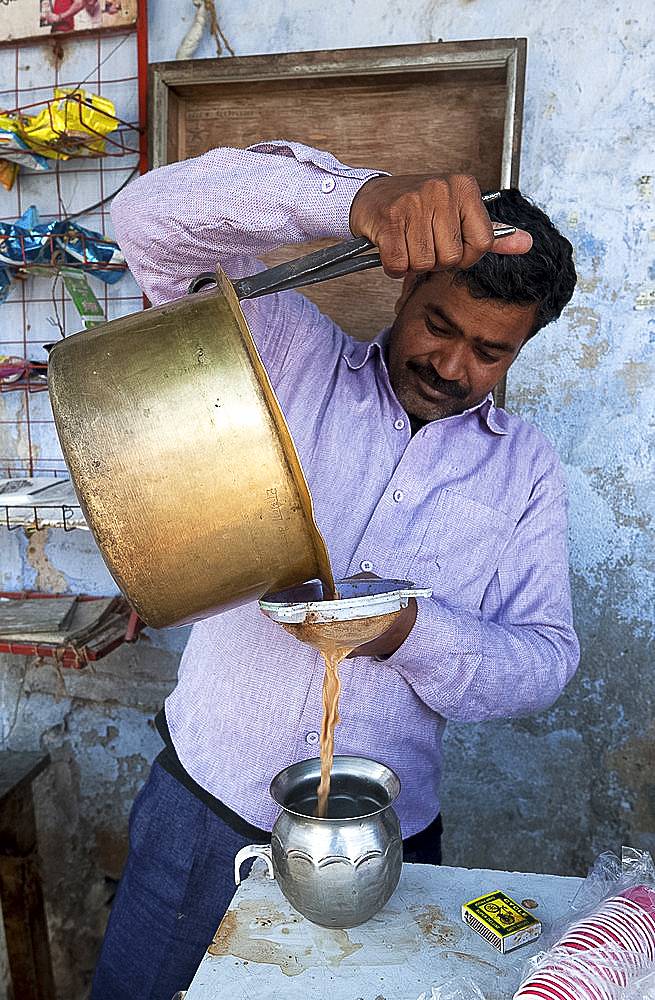 Chai wallah pouring chai from brass pan, using tongs, through sieve into tin vessel, in Diggi village market, Diggi, Rajasthan, India, Asia