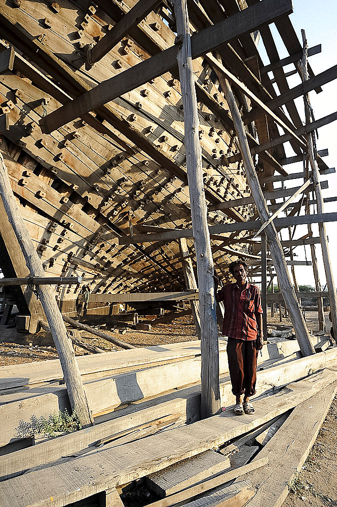 Boatbuilder beneath the hull of ocean going dhow under construction by hand from Sal wood, Rukmavati River, Mandvi, Gujarat, India, Asia